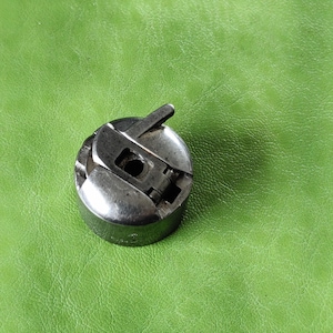 Może przedstawiać: Srebrna szpulka do maszyny do szycia z pokrywką na zawiasach i małą dźwignią na górze.