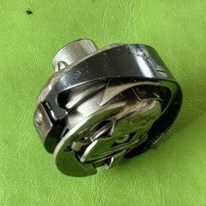 Pode incluir: Uma vista de perto de uma caixa de bobina de máquina de costura. A caixa da bobina é feita de metal brilhante e tem um anel externo preto. O fundo é verde texturizado sólido.