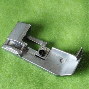 Può includere: Un piedino per macchina da cucire in argento con una vite e una piccola piastra metallica con il numero "20" su di essa.