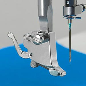 Può includere: Primo piano del piedino premistoffa e dell'ago di una macchina da cucire. Il piedino premistoffa è in metallo argentato lucido e ha una leva. L'ago è sottile e metallico, posizionato verticalmente. Lo sfondo è un mix di blu e grigio.