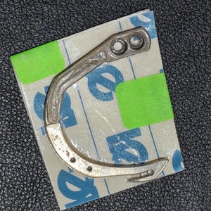 Pode incluir: Gancho de máquina de costura de metal prateado com forma curva e vários furos. O gancho está sobre um fundo branco com marcas azuis e verdes.
