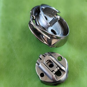 Könnte beinhalten: Zwei silberne Nähmaschinenspulen. Die größere Spule hat eine runde Form mit einem erhabenen, geschwungenen Design. Die kleinere Spule hat eine rechteckigere Form mit einem erhabenen, geschwungenen Design.