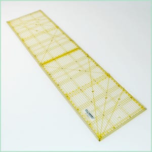 Puede incluir: Una regla de plástico transparente con marcas amarillas. La regla tiene 15 cm de ancho y 61 cm de largo. Tiene un patrón de cuadrícula con líneas diagonales y marcas para pulgadas y fracciones de pulgada. La regla está etiquetada con "OLFA" en la esquina inferior derecha.