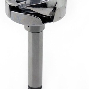 May include: A silver sewing machine bobbin case. The bobbin case has a cylindrical stem with three grooves near the base. The top section has a circular design with a curved metal piece and a central post.