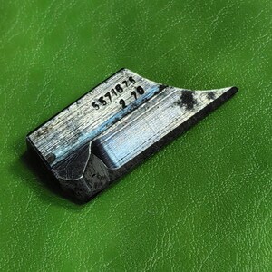 Può includere: Un piccolo utensile metallico angolato con i numeri "5571675 2 70" incisi sulla superficie. L'utensile ha un corpo rettangolare con un bordo affilato e appuntito. Il metallo ha un aspetto usurato e strutturato.