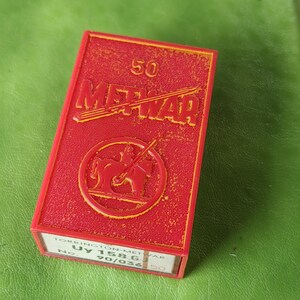 Pode incluir: Uma caixa vermelha com a palavra "METWAR" em letras amarelas. A caixa tem um emblema circular com um cavalo e um cavaleiro. O número "50" está no topo. A parte inferior da caixa tem o texto "TORRINGTON METWAR UY 1586 No. 90/036 50".