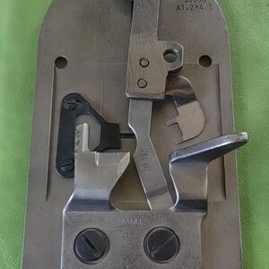 Può includere: Un pezzo di macchina da cucire in metallo argentato con una leva nera e diverse viti. Il pezzo è etichettato "20330 A1/2X4-5" e "20/114".