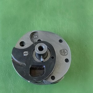 Könnte beinhalten: Ein silbernes und schwarzes Metallteil für eine Nähmaschine mit einem kleinen Loch in der Mitte und einer kreisförmigen Rille am Rand. Das Teil hat einen kleinen Metallstift, der aus dem mittleren Loch herausragt. Das Teil hat eine kleine Metallschraube an der Seite. Das Teil hat ein kleines Metallschild mit dem Text "Made in Germany" darauf.