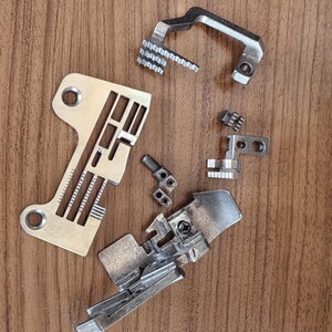 May include: Various silver sewing machine components, including presser feet, feed dogs, and a needle plate, are displayed on a wooden surface. The metallic parts showcase detailed designs, indicating precision engineering for fabric manipulation.