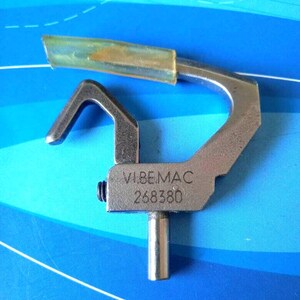 Pode incluir: Um pé de máquina de costura de metal prateado com um guia de plástico transparente. O pé tem o texto "VIBEMAC 268380" gravado.