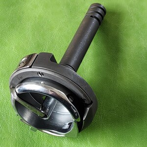 May include: A silver and black sewing machine bobbin case with a metal hook and a black metal shaft.