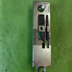 Op de afbeelding: Een zilverkleurig naaimachineonderdeel met de tekst "PFAFF" en "48025C/D". Het onderdeel heeft een rechthoekige vorm met meerdere groeven en twee ronde gaten. De achtergrond is effen groen. Metrische maten.