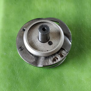 Könnte beinhalten: Eine silberne Nähmaschinenspule mit einem schwarzen Metallsockel und einer kleinen Schraube an der Seite. Die Spule hat eine Rille am Rand und ein kleines Loch in der Mitte.