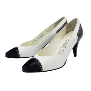 May include: A pair of white leather pumps with black cap toes and heel accents. The shoes have a pointed toe design and are embellished with small, decorative studs. The heels are approximately 3 inches high.