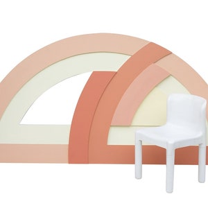 May include: A white plastic chair sits in front of a colorful, abstract wall made of overlapping semi-circles in shades of pink and cream.