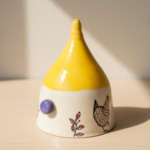 May include: A ceramic decorative piece with a yellow top and a white base. The base features a purple knob, a chicken illustration, and a red and black leaf design. The piece is on a wooden surface.