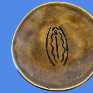 May include: A brown ceramic plate with a black line drawing of a vulva. The plate has a glossy finish and a slightly uneven surface. The background is a solid blue.