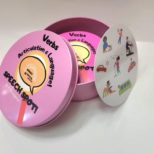 May include: A pink, round tin labeled "Speech Spot!" contains cards for articulation and language practice. The tin's lid features the words "Verbs" and "Articulation & Language!" A circular card with illustrations of various actions is visible.