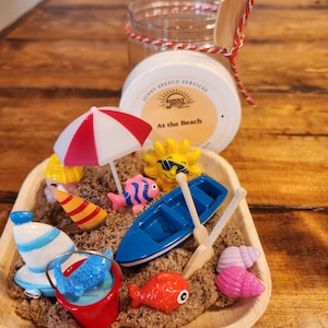 May include: A miniature beach scene with a red and white umbrella, a blue boat with oars, a yellow sun with sunglasses, a red and blue striped beach towel, a blue bucket with a blue fish, a pink seashell, and a red fish. The scene is set in a brown tray with sand.