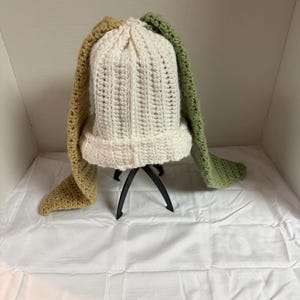 May include: A cream-colored crocheted beanie featuring two long, dangling ear-like attachments. One attachment is a muted mustard yellow, while the other is a sage green. The hat has a ribbed texture and a folded brim, crafted from yarn.