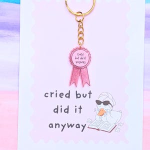 May include: A gold-toned metal keychain featuring a pink acrylic ribbon charm with the text Cried but did it anyway. The charm is attached to a display card with the same text and an illustration of a duck wearing sunglasses and a towel.