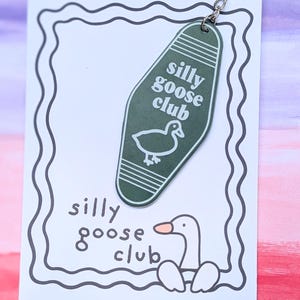 May include: A dark green motel-style keychain featuring the text "silly goose club" and a white goose illustration. It is attached to a white display card with a wavy border, the text "silly goose club," and a cartoon goose graphic.