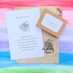 May include: A silver-toned metal charm featuring multiple small spoon shapes attached to a heart-shaped ring, accompanied by a white informational card titled The Spoonie Tool and a small brown cardboard box, all resting on a tan burlap pouch.