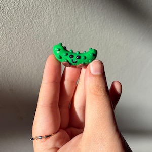 May include: A small, bright green, bumpy pickle figurine with a glossy finish. The pickle has two black dot eyes, a small black curved smile, and two tiny brown feet at the base. The surface of the pickle is textured with small raised bumps.