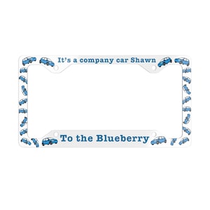 May include: White license plate frame with blue car illustrations and the text "It's a company car Shawn" and "To the Blueberry". The frame has a rectangular shape with rounded corners and mounting holes.