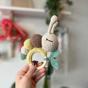 Puede incluir: Un sonajero de bebé hecho a mano de ganchillo con forma de oruga con cabeza de conejo. El sonajero tiene un anillo de madera y está hecho con hilo beige, marrón, amarillo y verde. El conejito tiene los ojos cerrados.