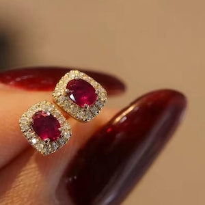 Może przedstawiać: Para złotych kolczyków sztyftów. Każdy kolczyk ma owalny, czerwony kamień otoczony otoczką małych diamentów. Klasyczny, elegancki design dodaje luksusu. Idealne na różne okazje.