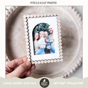May include: A white wooden picture frame with a scalloped edge and an arched opening. The frame is designed to hold a 2.5 x 3.5 inch photo. The frame is shown in a hand holding it.