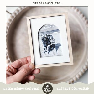 May include: A laser-cut wooden frame with an arched opening, designed to fit a 2.5 x 3.5 inch photo. The frame is light brown and has a white mat. The frame is held in a hand, and a black and white photo of three people on a beach is visible inside the frame.