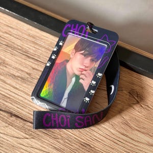 May include: A black plastic ID card holder featuring a purple and white cat-themed design with paw prints and the text CHOI SAN. The holder contains a holographic photo card of a person and is attached to a black lanyard with purple text.