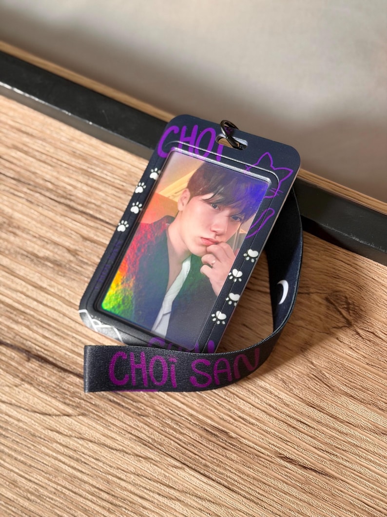 May include: A black plastic ID card holder featuring a purple and white cat-themed design with paw prints and the text CHOI SAN. The holder contains a holographic photo card of a person and is attached to a black lanyard with purple text.