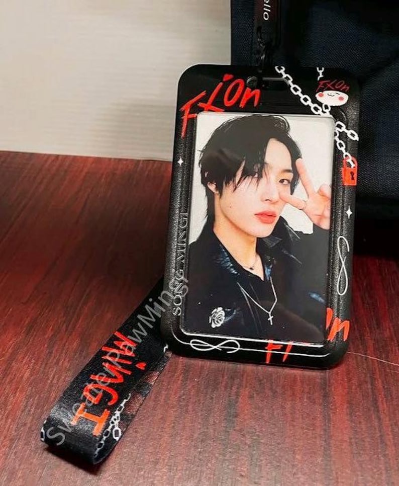 May include: A black plastic ID card holder featuring a graphic of a person making a peace sign. The holder is decorated with red text reading "Fix On," white chain patterns, and a small red padlock. A matching black lanyard with red text is attached.