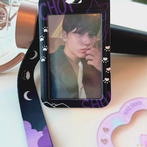 May include: A black plastic ID card holder featuring a portrait of a person, attached to a black lanyard with purple cloud, moon, and cat paw print patterns. The holder has the text CHOI printed in purple and includes a small black clip at the top.