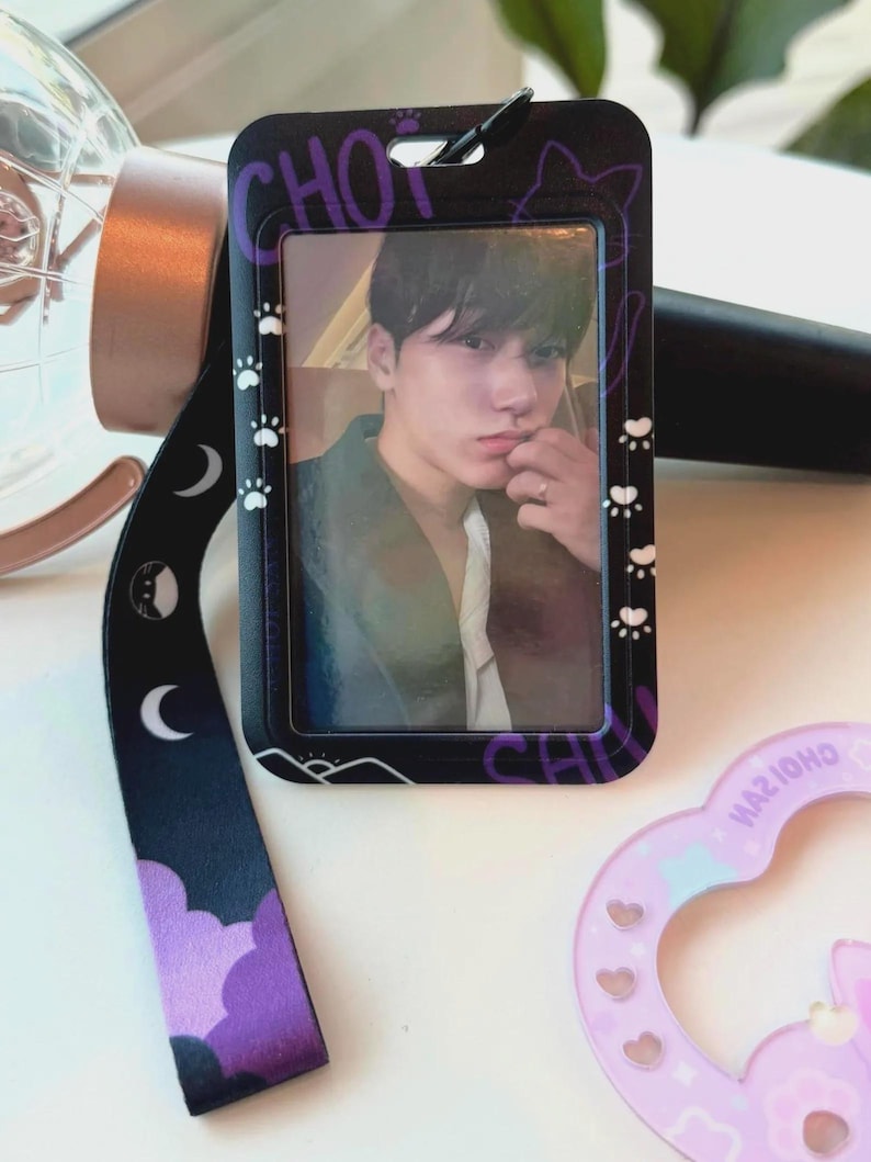May include: A black plastic ID card holder featuring a portrait of a person, attached to a black lanyard with purple cloud, moon, and cat paw print patterns. The holder has the text CHOI printed in purple and includes a small black clip at the top.