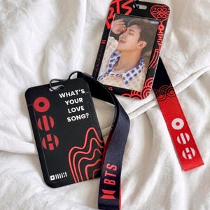 May include: Two black and red plastic badge holders with matching lanyards. One holder features a photo of a person in a yellow and blue plaid shirt, while the other displays the text "WHAT'S YOUR LOVE SONG?" and the word "BTS" on the lanyard.
