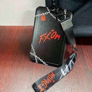 May include: A black rectangular ID badge holder featuring a white chain link pattern and a red padlock icon. The text "FXOn" is printed in red. A matching black lanyard with white chain links and red text is attached via a black metal clip.
