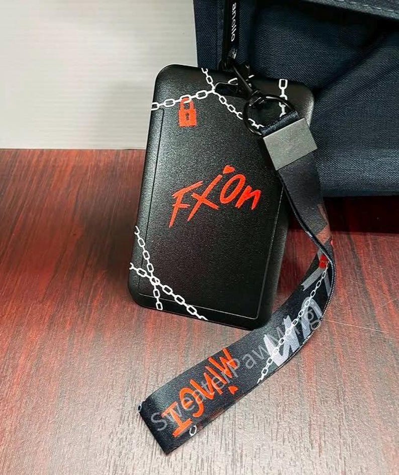 May include: A black rectangular ID badge holder featuring a white chain link pattern and a red padlock icon. The text "FXOn" is printed in red. A matching black lanyard with white chain links and red text is attached via a black metal clip.