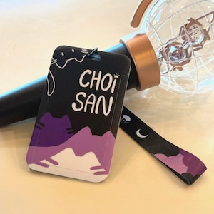 May include: A black and purple rectangular tag with the text CHOI SAN printed in white, featuring a cat illustration. The tag is attached to a black wrist strap with moon and cat icons, resting near a clear globe-shaped light stick with a rose gold base.