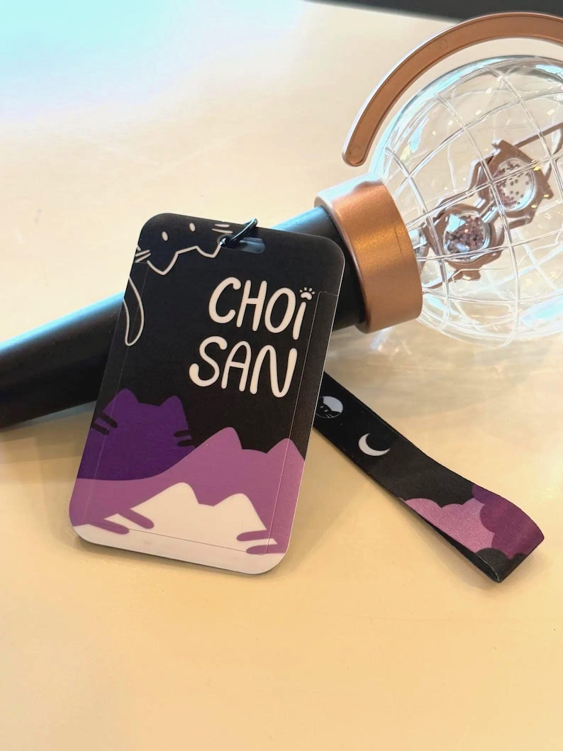 May include: A black and purple rectangular tag with the text CHOI SAN printed in white, featuring a cat illustration. The tag is attached to a black wrist strap with moon and cat icons, resting near a clear globe-shaped light stick with a rose gold base.