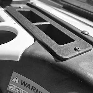 May include: Close-up of a black rectangular frame with two openings, secured with screws. The frame sits on a dark surface, with a white curved edge visible on the left. A warning label is partially visible.