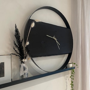 Könnte beinhalten: Eine moderne schwarze Wanduhr mit goldenen Zeigern an einer weißen Wand. Die Uhr hat einen kreisförmigen Metallrahmen und ein rechteckiges schwarzes Zifferblatt. Ein Regal darunter beherbergt eine Vase mit Trockenblumen und eine kleine Topfpflanze.