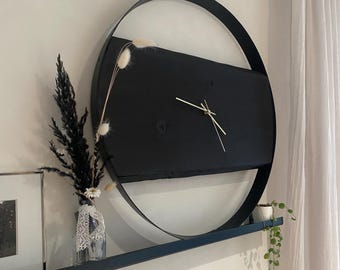 May include: A modern black wall clock with gold hands, set against a white wall. The clock features a circular metal frame and a rectangular black face. A shelf below holds a vase of dried flowers and a small potted plant.