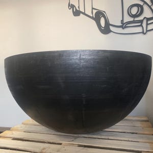 May include: A large, black, metal bowl sits on a wooden pallet. The bowl is round and has a smooth, slightly textured surface.