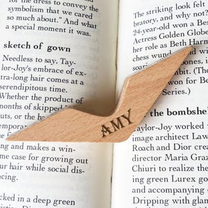 Puede incluir: Un marcap&aacute;ginas de madera clara con un dise&ntilde;o angular, colocado sobre un libro abierto. El marcap&aacute;ginas tiene grabado el nombre "AMY". Un accesorio elegante y pr&aacute;ctico para los amantes de la lectura.