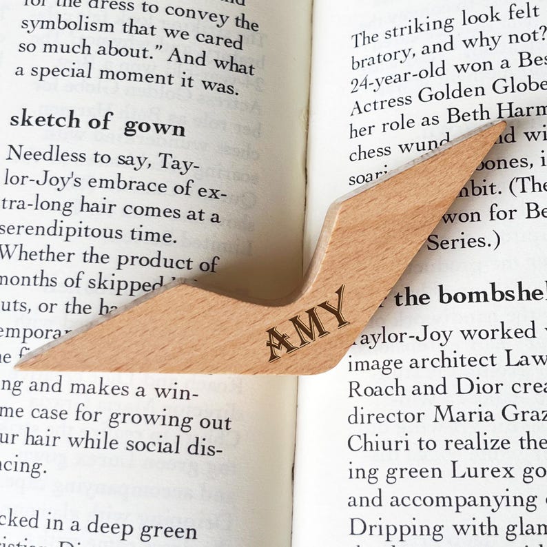 Puede incluir: Un marcap&aacute;ginas de madera clara con un dise&ntilde;o angular, colocado sobre un libro abierto. El marcap&aacute;ginas tiene grabado el nombre "AMY". Un accesorio elegante y pr&aacute;ctico para los amantes de la lectura.
