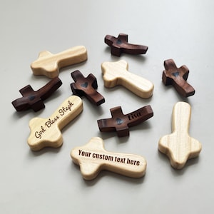 May include: A collection of wooden crosses in various shades of brown and natural wood. Some crosses have engraved text, including "God Bless Steph", "Lisa", and "Your custom text here". Several crosses feature a small heart design.